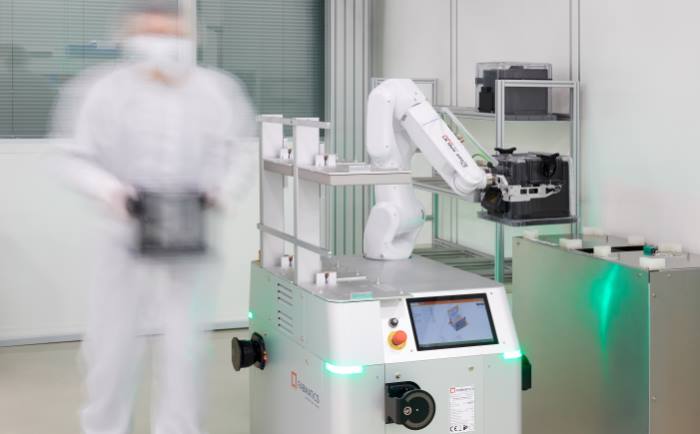 Operator in cleanroom clothing carries wafer FOUP, mobile robot handles FOUP with wafers on cleanroom shelf to the right of it