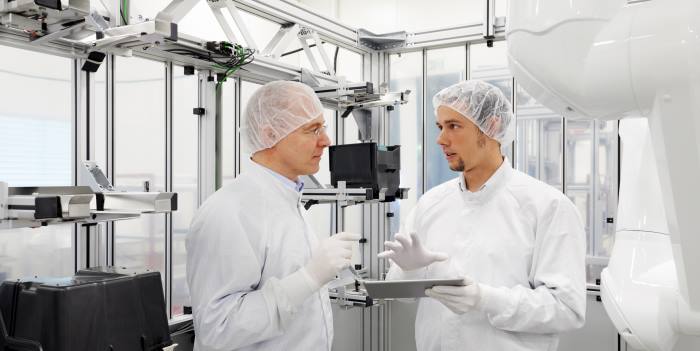 Two men in cleanroom gowns are talking on a tablet.